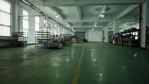 A wide shot of the interior of a sparsely filled factory. Workers are sitting at a long table near the windows, surrounded by shelves. Behind them, some distance away, are shelves with boxes stacked on them.