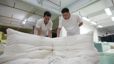 Two men in white shirts, the bosses of a sofa factory, bend over to examine a white sofa in a factory.