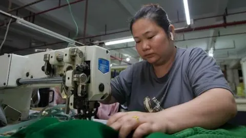 A woman wearing a grey t-shirt at a sofa factory sits in front of a sewing machine. She is wearing white earbuds and holding a large piece of green fabric at the table.