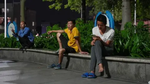 Three unemployed men sit on a ledge in a park with some distance between each of them. One is wearing a blue shirt and squatting, looking at his phone; another wearing a bright yellow shirt is leaning on his hand with one foot on the ledge, looking at the camera; beside him is another man in a white shirt with his legs crossed and looking down at his phone.