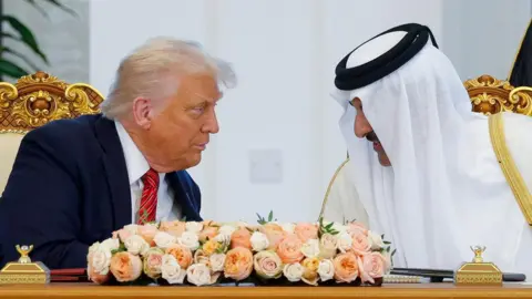 Reuters Trump and Al Thani lean towards each other in conversation behind an ornate gold desk with a bunch of white and orange and pink flowers on it 
