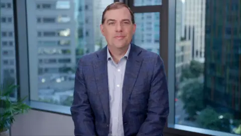 BBC A medium close up of Matt Garman, chief executive Amazon Web Services, sat facing the camera in front of a window of an office building with the city outside visible. He is smiling slightly and wearing a blue suit and a shirt without a tie.