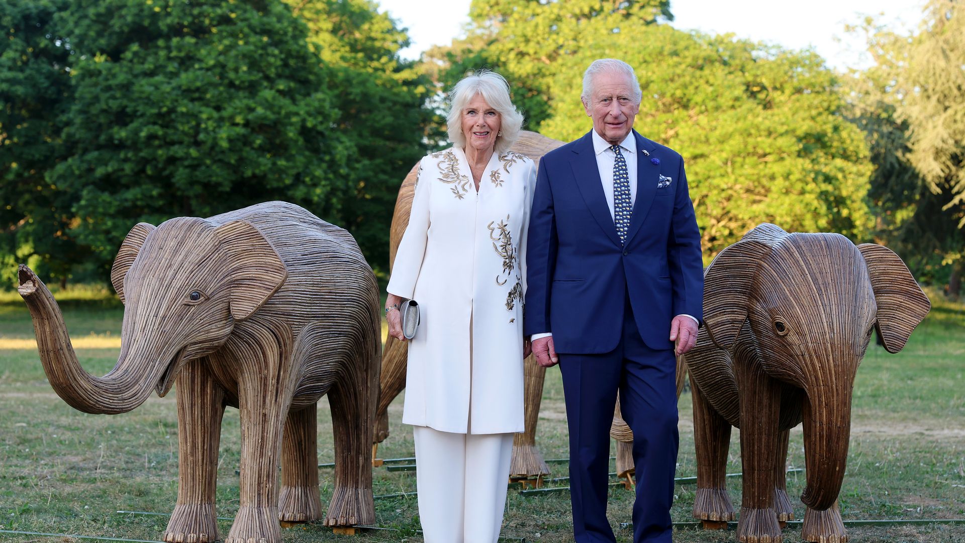 Queen Camilla and King Charles standing in between wooden elephants