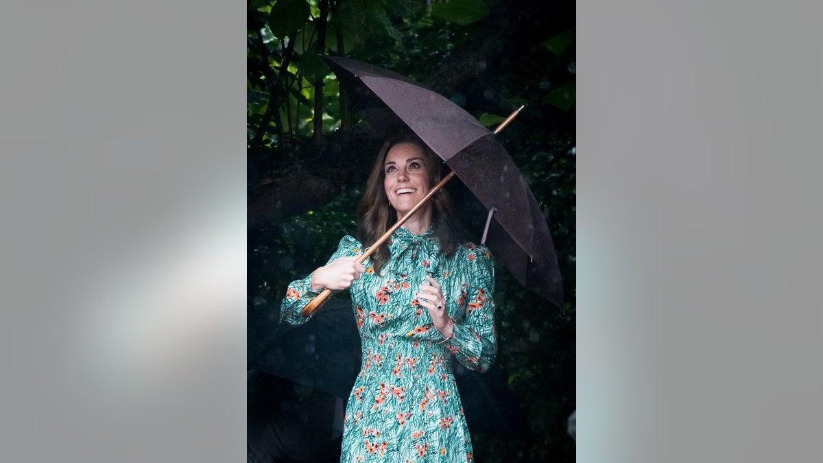 Kate Middleton wearing a green floral dress holding an umbrella in the rain and smiling.