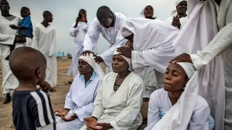 NurPhoto/Getty Images Three female apostolic church members sit with their legs in front of them and their palms upturned on their laps to receive blessings from fellow congregants who place hands on their heads. They are gathered outside in a rocky area in Harare and everyone is dressed in white expect for a child looking on and another child being held by a man in the background.