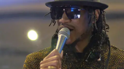 Jah Prayzah, wears shades, a black hat and in a yellow tweed-like top, as he sings holding a microphone in a BBC studio in 2021