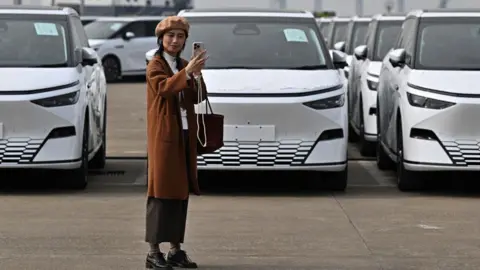 Getty Images A woman takes a selfie in front X9 electric vehicles by Chinese EV manufacturer XPeng, are loaded on ship of the NYK line during a ceremony in the Port of Guangzhou, China's southern Guangdong province, on 22 February, 2025.