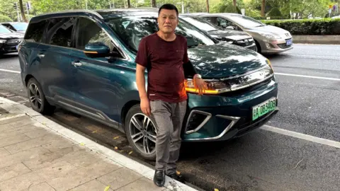 Annabelle Liang Lu Yunfeng, a private hire driver, stands in front of his teal Dayun electric car that is parked along a road in Guangzhou in China. He is wearing a maroon t-shirt, striped grey pants and black shoes.