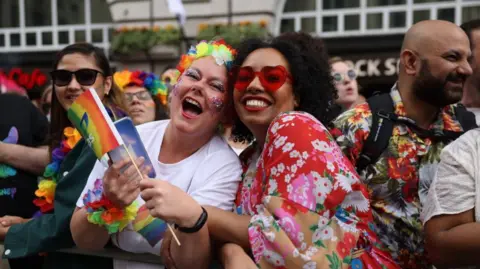Getty Images A picture focused on two attendees of a previous Pride in London event. On the left, a woman with glitter on her face and with flowery decorations around her head and arms laughs at the camera. On the right, another woman smiles at the camera with red-heart sunglasses on and a red flowery dress.