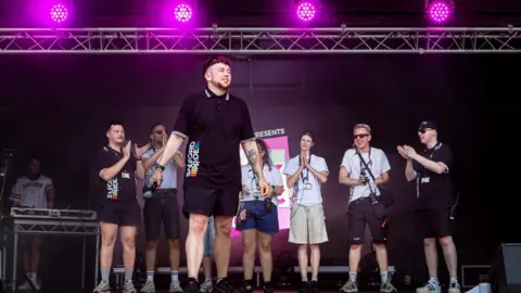 OhMyMedia / Olly Rigby Reece stands on stage at the Pink Picnic event in Salford. With pink stage lights above him, he is holding a microphone by his side and has colleagues applauding behind him. His is wearing a black polo top with Salford Pride written on the side and blackshorts.