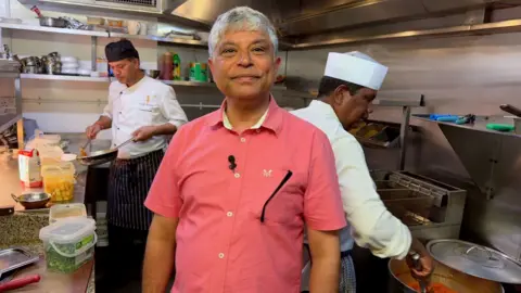 Sanjay Jha is standing in the kitchen with two staff behind who are preparing food.