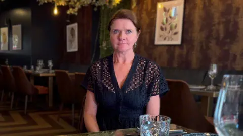 Kate is sitting at the table of an empty restaurant in London. She has a glass in front of her at the table and there is a row of empty seats at a table behind. She has brown hair and is wearing a navy blouse