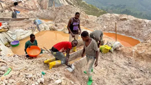 Hassan Lali / BBC A group of five miners, wearing wellingtons, work at station cleaning the rocks brought up to the surface. They are standing next to a pool of orange, brown water used in the process. Behind them can be seen the Masisi Hill.