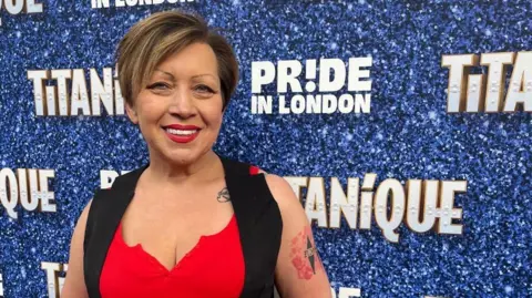 Dee Llewellyn A head and shoulders shot of Dee smiling into the camera, in front of a Pride in London branding board on a red carpet. She is wearing a black vest over a red top and has a red and black tattoo on her upper arm.