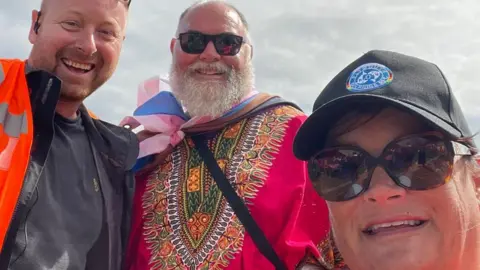 Alex MacDonald Alex is pictured with two other volunteers outside at a previous Plymouth Pride event. All three are smiling at the camera, while Alex wears an orange hi-vis jacket over a dark top.