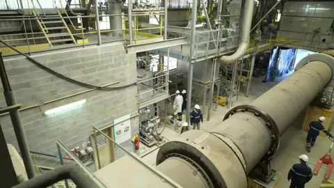 Workers in blue safety jackets and white hard hats work around a giant kiln at Solvay's rare earths processing facility in La Rochelle