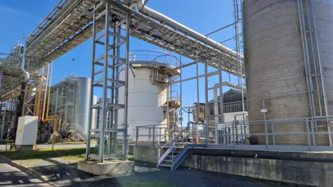 BBC Storage tanks and overhead pipes amid a bright blue sky at Solvay's rare earths processing facility in La Rochelle, western France