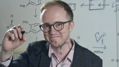 University of Edinburgh Smiling Professor Stephen Wallace writes chemical formulae on a see-through display board.
