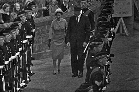 Black and white photo of Queen Elizabeth II and an unidentified man walking between lines of guardsmen.