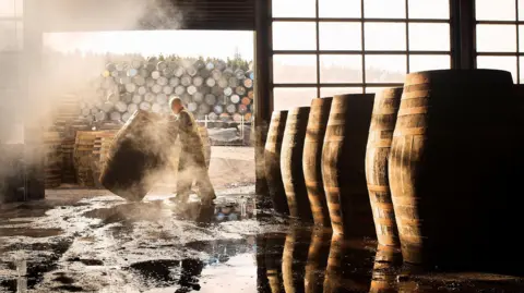 Getty Images A man moves a whisky barrel in a warehouse. There are dozens of barrels in the background (outside of the warehouse). Inside the warehouse there are six large barrels. 