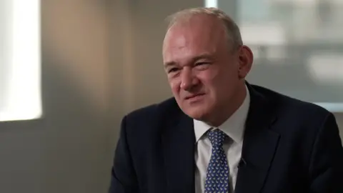BBC Sir Ed Davey looks to the left of the camera. He is wearing a dark suit and blue and white tie. 
