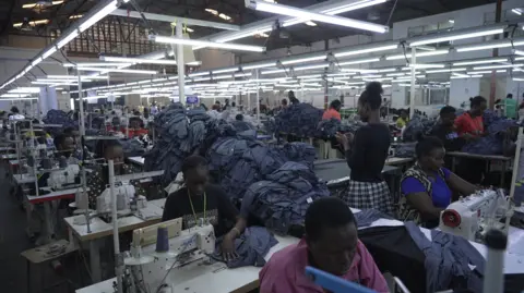 Hassan Lali / BBC A wide shot of a busy factory floor of the garment factory. Workers at sewing machines and piles of finished items can be seen.