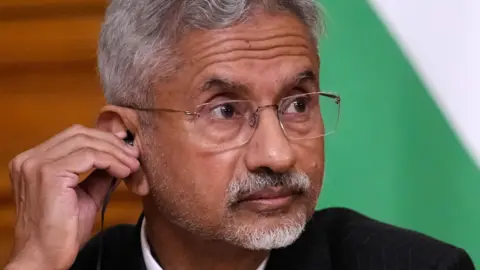 EPA Subrahmanyam Jaishankar with white hair and goatee, glasses, and dark suit raises his eyebrows as he puts a black earbud in his ear, in front of a green and white background