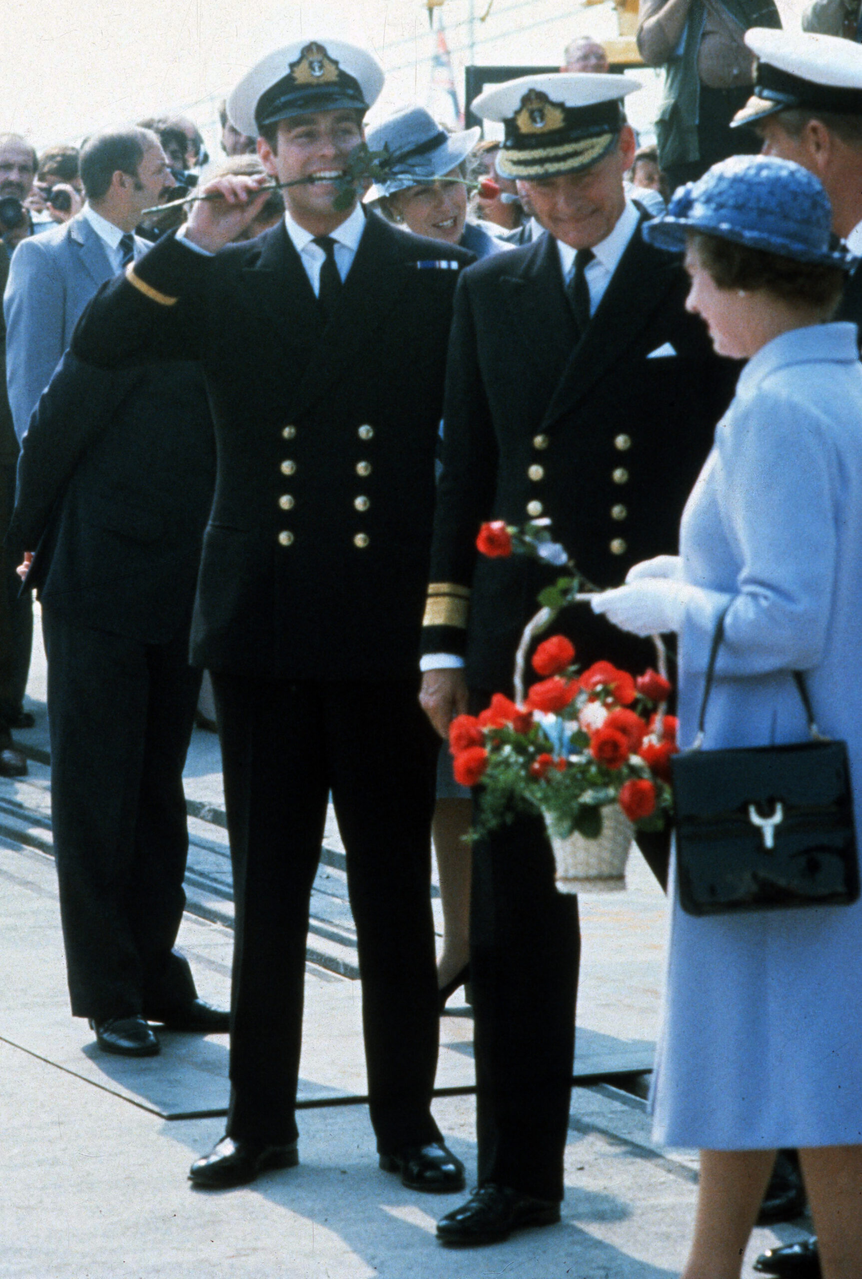 Prince Andrew holds a rose in his mouth, with Queen Elizabeth II standing to his right holding a basket of roses.