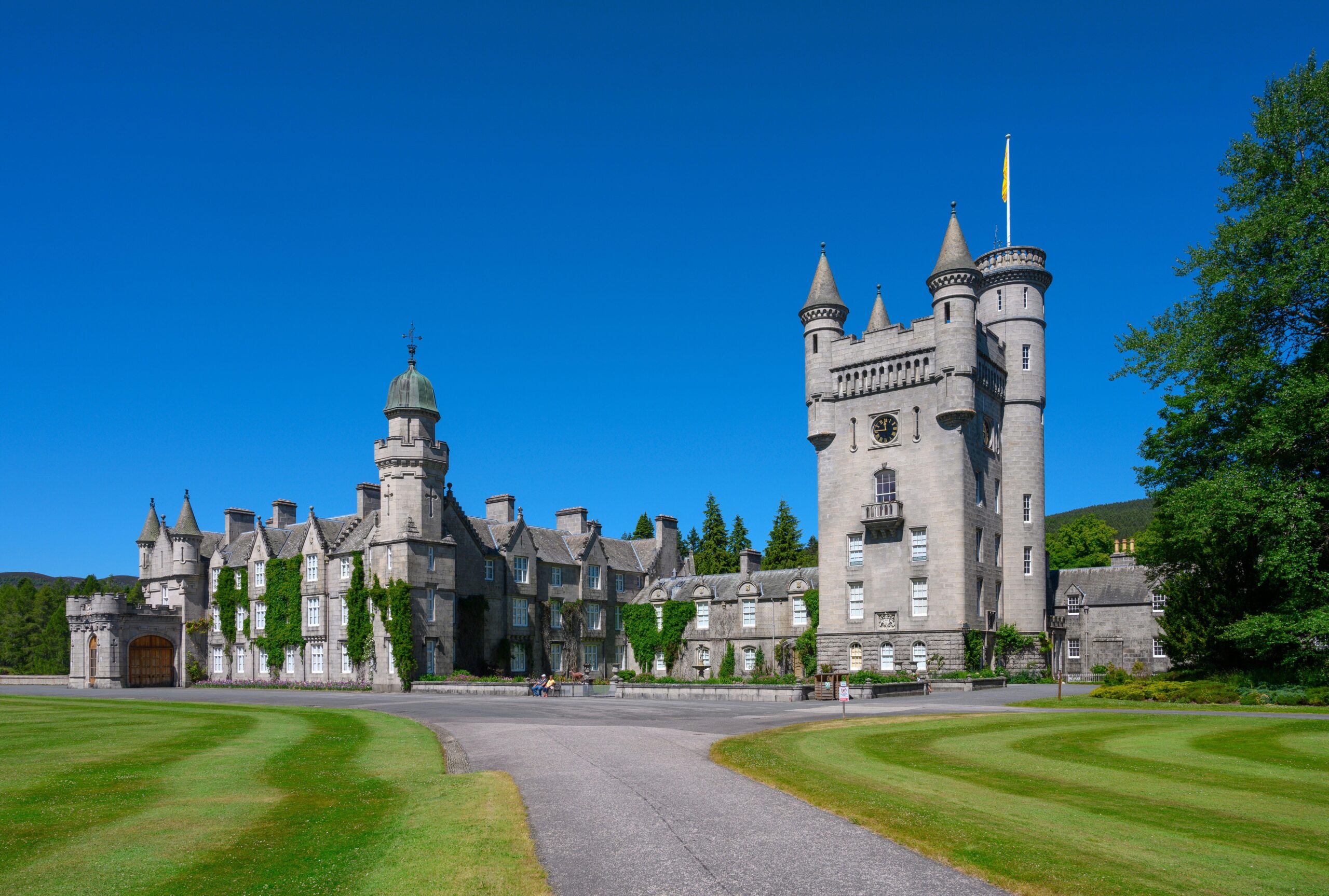 Balmoral Castle, with its towers and spires, surrounded by green lawns and trees under a clear blue sky.