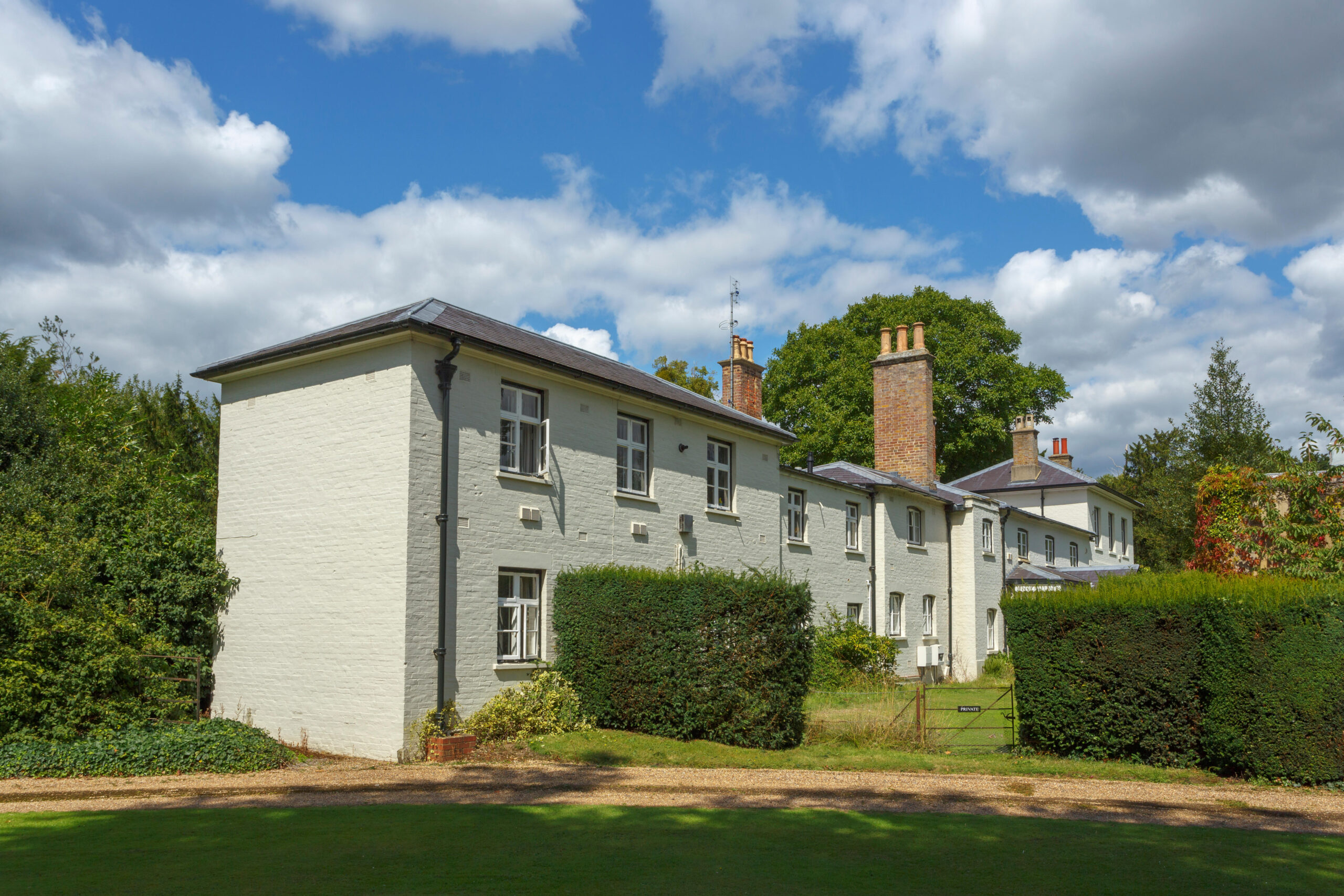 Frogmore Cottage, a white brick building with multiple windows and chimneys, surrounded by green hedges and trees, under a blue sky with white clouds.