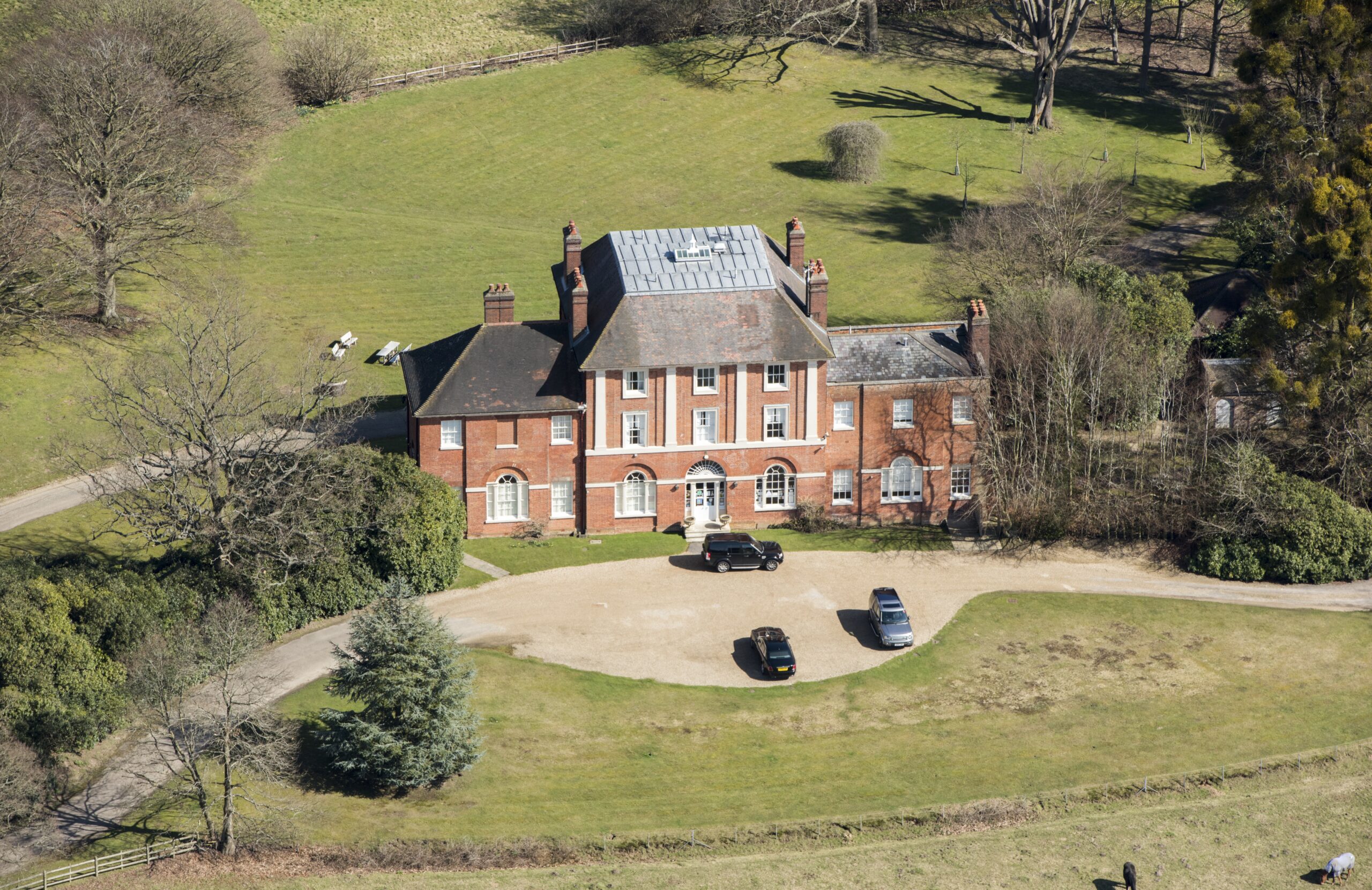 Aerial view of Forest Lodge, a large brick house with a gravel driveway and cars, surrounded by trees and green fields.