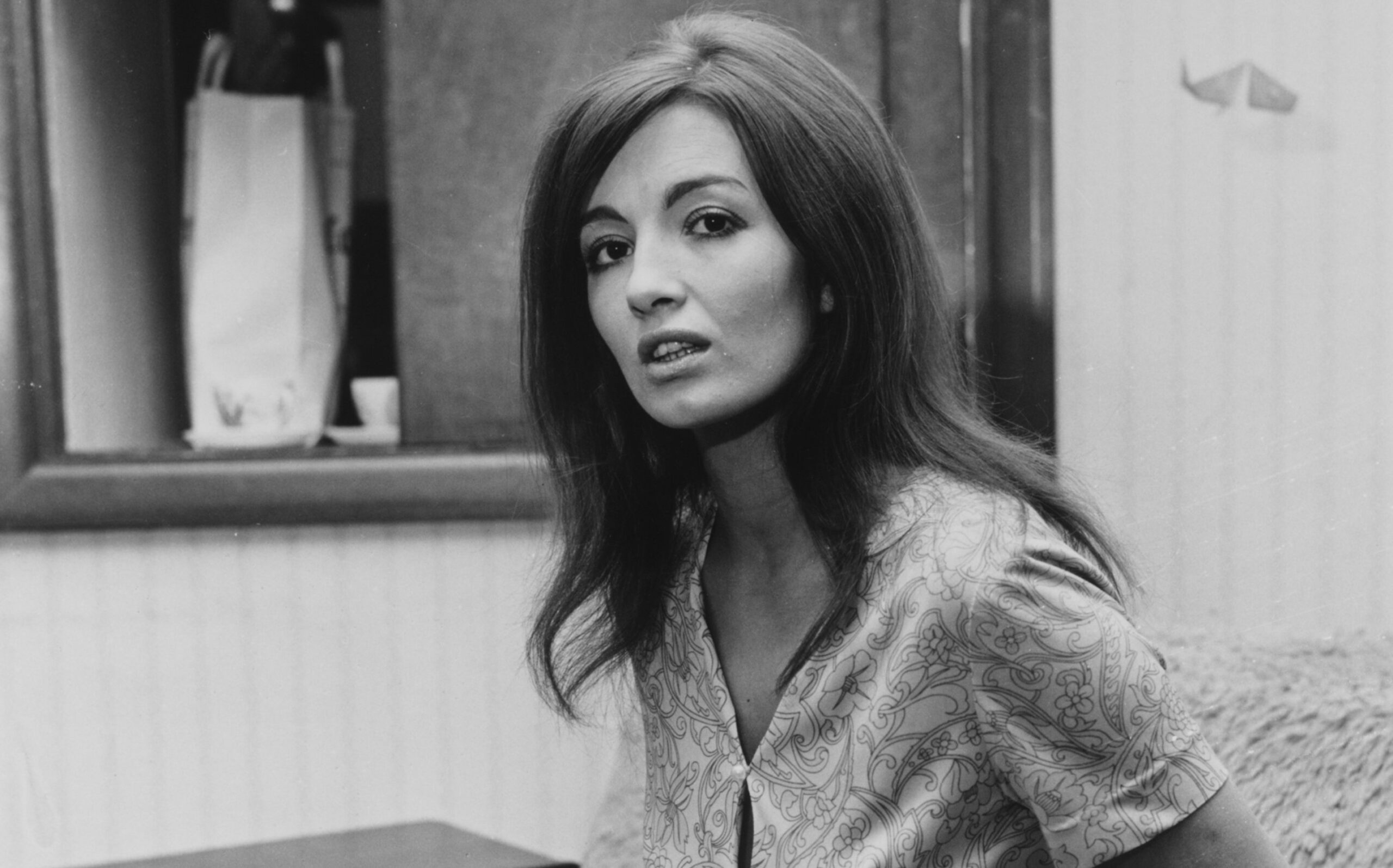 Christine Keeler wearing a patterned mini-dress and sitting on a fur-covered couch.
