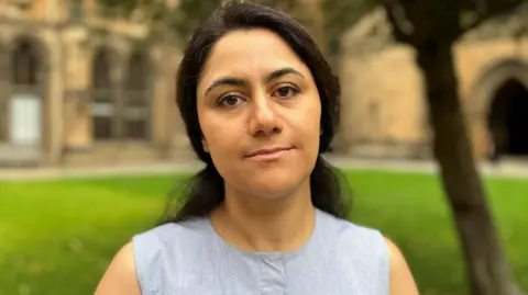 Prof Basiri looks at the camera. She is outside in the quadrangle at Glasgow university. The university buildings can be seen in the background. She has dark hair and is wearing a blue sleeveless tunic top.