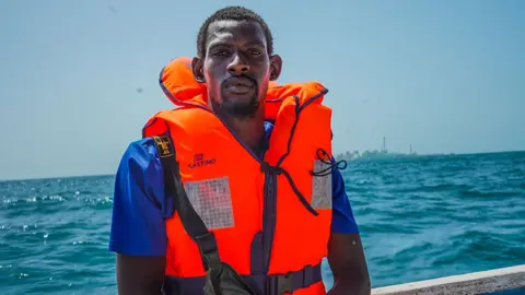 Michel Mvondo / BBC A shot of a man from the waist up. He is wearing a bright orange life jacket over a purple T-shirt. The edge of a canoe and the sea can be seen behind him.