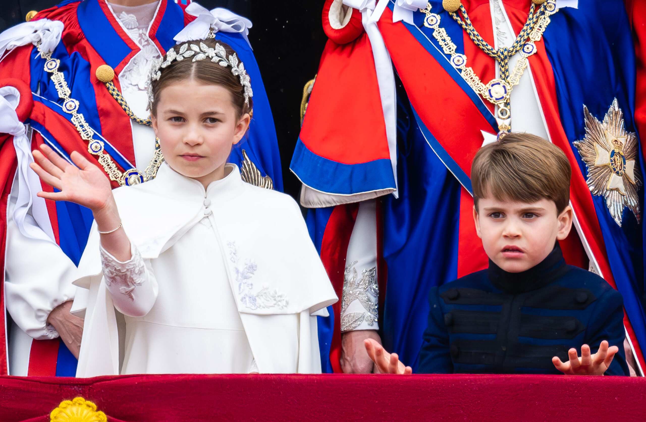 Princess Charlotte waving at the coronation in a white cape standing next to Prince Louis, gesturing with his palms