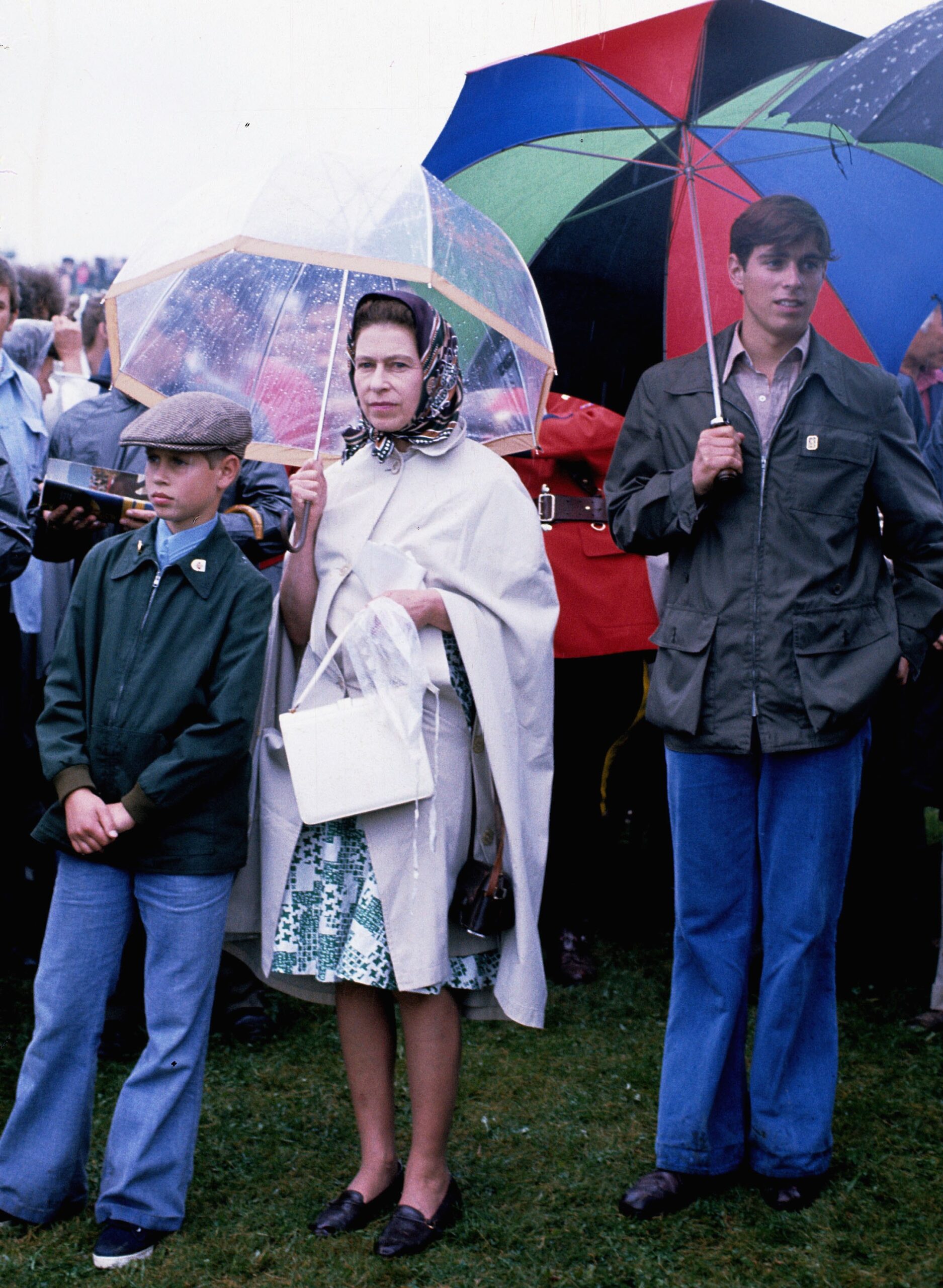 A teenage Prince Andrew holding an umbrella next to Queen Elizabeth and Prince Edward, also standing under an umbrella