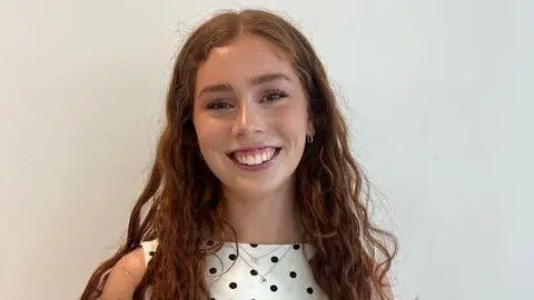 Grace Warren Grace has long brown hair and is wearing a polka dot top. She is standing in front of a white background and smiling.