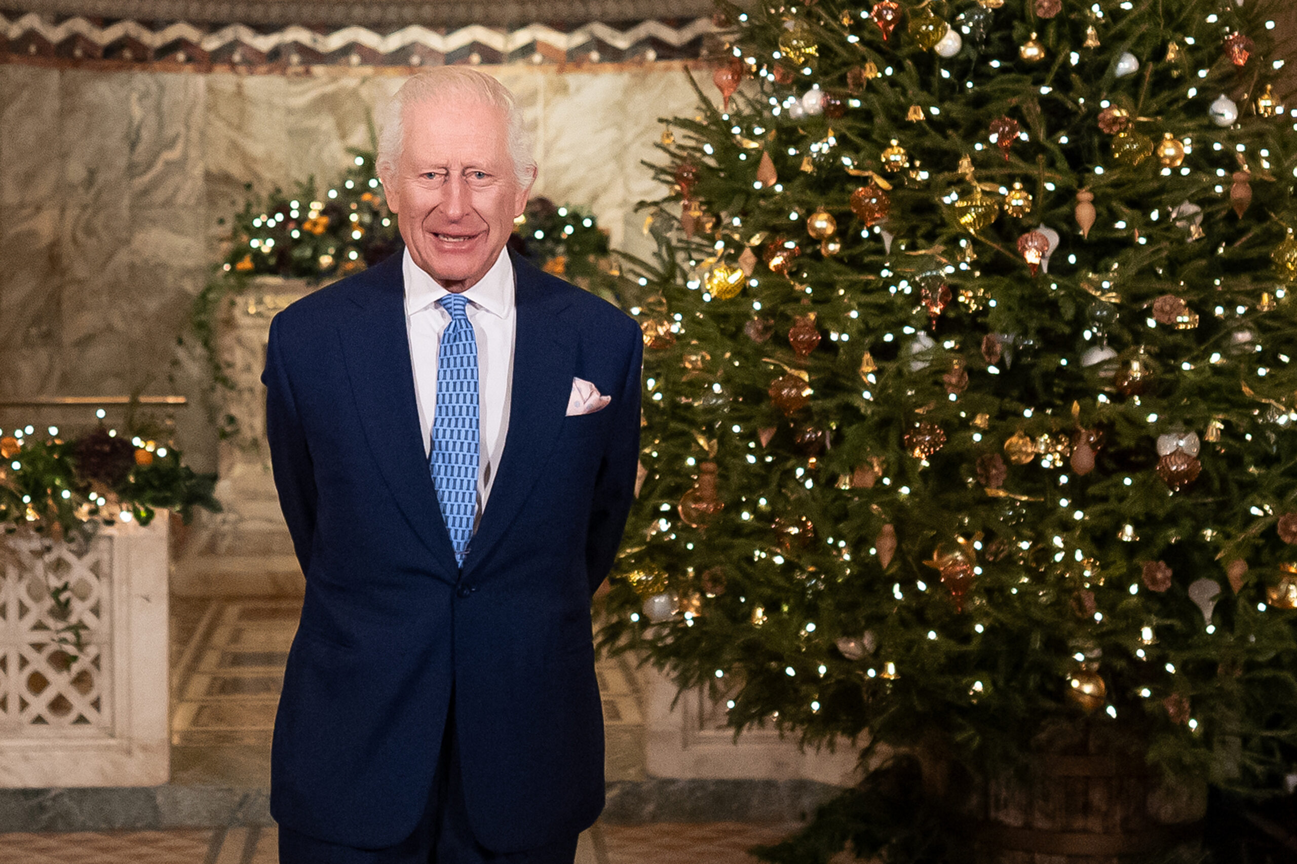 King Charles talking in front of a lit Christmas tree wearing a blue suit and tie
