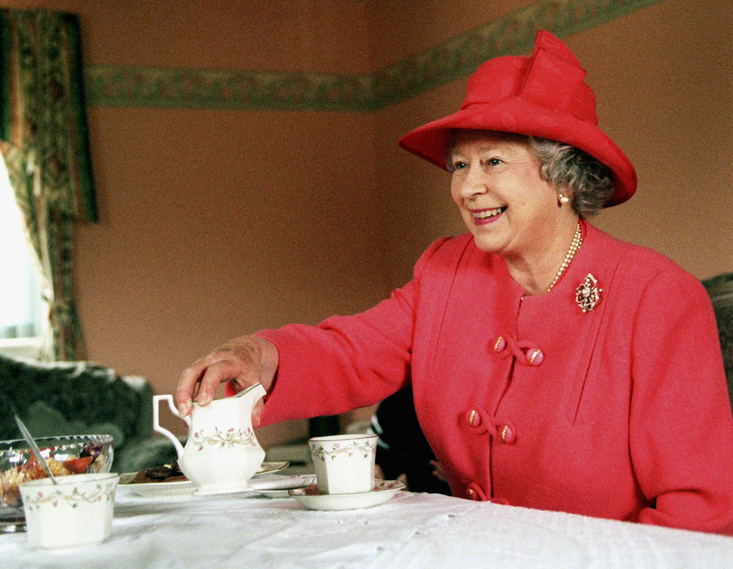 Queen Elizabeth wearing a pink suit drinking tea at a table