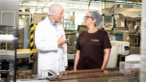 Chocolats Camille Bloch Daniel Bloch in a white lab coat and hair net stands with a woman in a black Camille Bloch t-shirt and hair net before a tray of chocolate bars in a factory