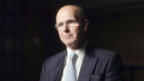 Sir Jeremy Fleming is dressed in a dark navy suit jacket with structured shoulders, a crisp white dress shirt, and a pale blue textured tie. He is seated in a dimly lit indoor setting with soft shadows and a dark, neutral background. The lighting highlights the suit fabric and tie texture, suggesting a formal or professional environment.