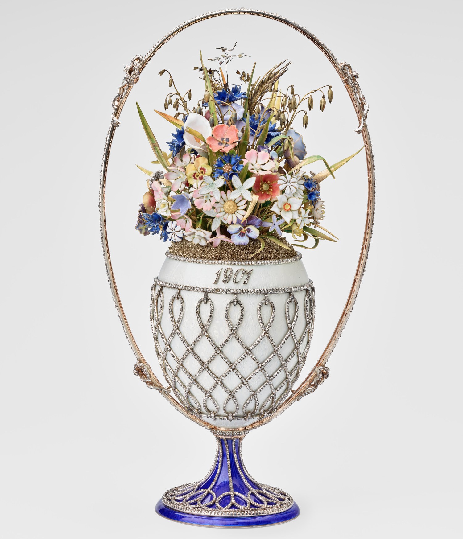 A jeweled Fabergé Basket of Flowers egg with a white basket base, a blue base, and a golden handle encasing a floral bouquet.