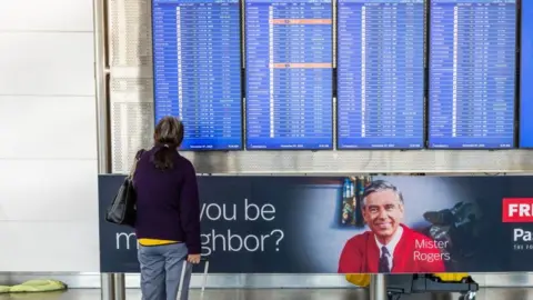 EPA/ Shutterstock A traveler dragging a suitcase on wheels looks at a flight arrival and departure display in an airport