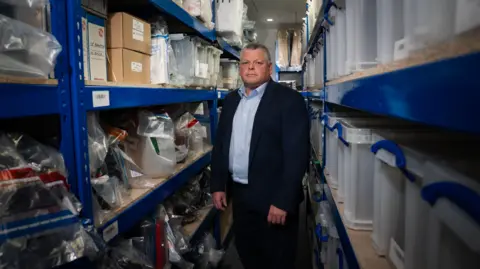 BBC Paul Curtis stands facing the camera in the aisle of the evidence room, piled high with luxury goods seized from financial fraudsters