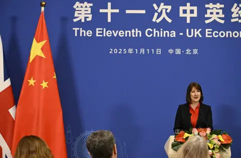 Getty Images A podium decorated with a floral arrangement stands in front of a blue backdrop displaying Chinese and English text that reads “The Eleventh China-UK Economic…” along with the date  and location in Chinese letters. To the left of the podium are two flags: the national flags of China and the United Kingdom. Several people are seated in the foreground, facing the podium.