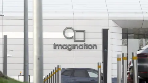 Exterior view of a modern white building with the Imagination Technologies logo on the wall, featuring a stylized circular and square design above the word ‘Imagination.’ Several bollards line the foreground, and parked cars are visible near the entrance.