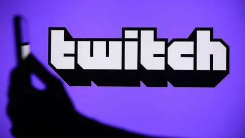 Getty Images Logo of Twitch platform on a purple background with a blurred hand holding a mobile phone 