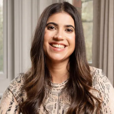 Rii Schroer A woman in a patterned brown and black top with long brown, curly hair sits in front of a window