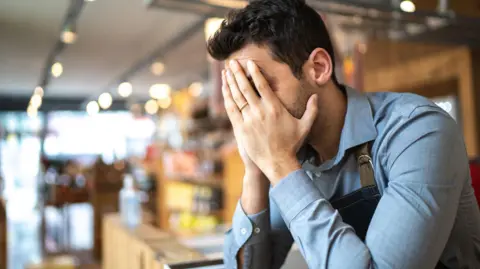 Getty Images A man wearing a blue shirt holds his head in his hands whilst standing at the serving bar. The cafe is visible but blurred in the background. 