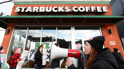 Reuters A young female union leader in a black and red woollen hat is standing among other picketers outside a Starbucks. Some of the other picketers in the background are holding signs whose message is not visible in the image. The female union member in the foreground is holding a megaphone as if she is about to start speaking into it.